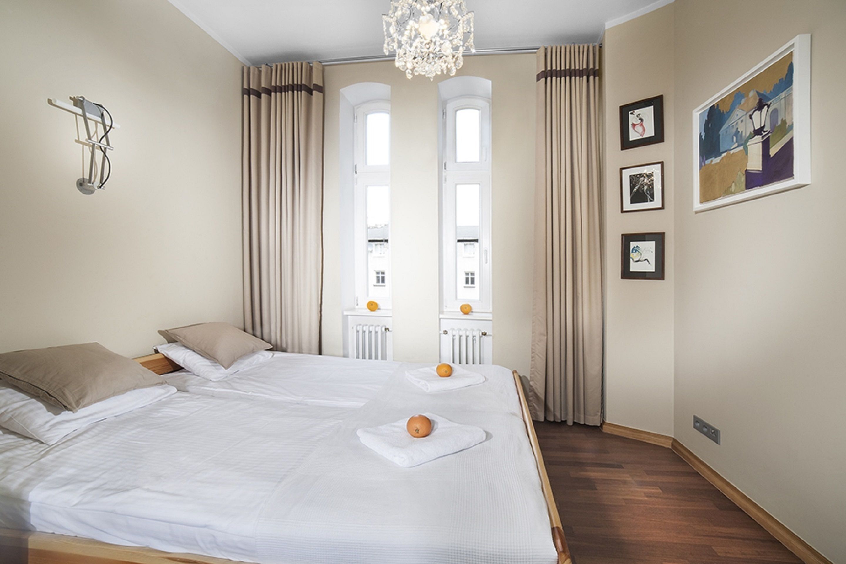 A hotel room with two beds, a chandelier, large windows with curtains, and framed artwork on the wall.