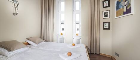 A hotel room with two beds, a chandelier, large windows with curtains, and framed artwork on the wall.