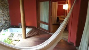 A hammock in a room with a stone wall, a wooden floor, and a view of a bathroom with a shower and a sink. - Pousada dos Ponteiros (São Miguel do Gostoso)