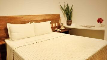 Una cama bien hecha con cabecera de madera, una mesita de noche con lámpara, una planta en maceta y un pequeño estante con libros y un jarrón de flores.