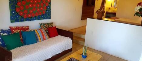 Una sala de estar con un sofá, cojines coloridos, una mesa de centro con una revista y un cuadro grande con motivos florales en la pared.