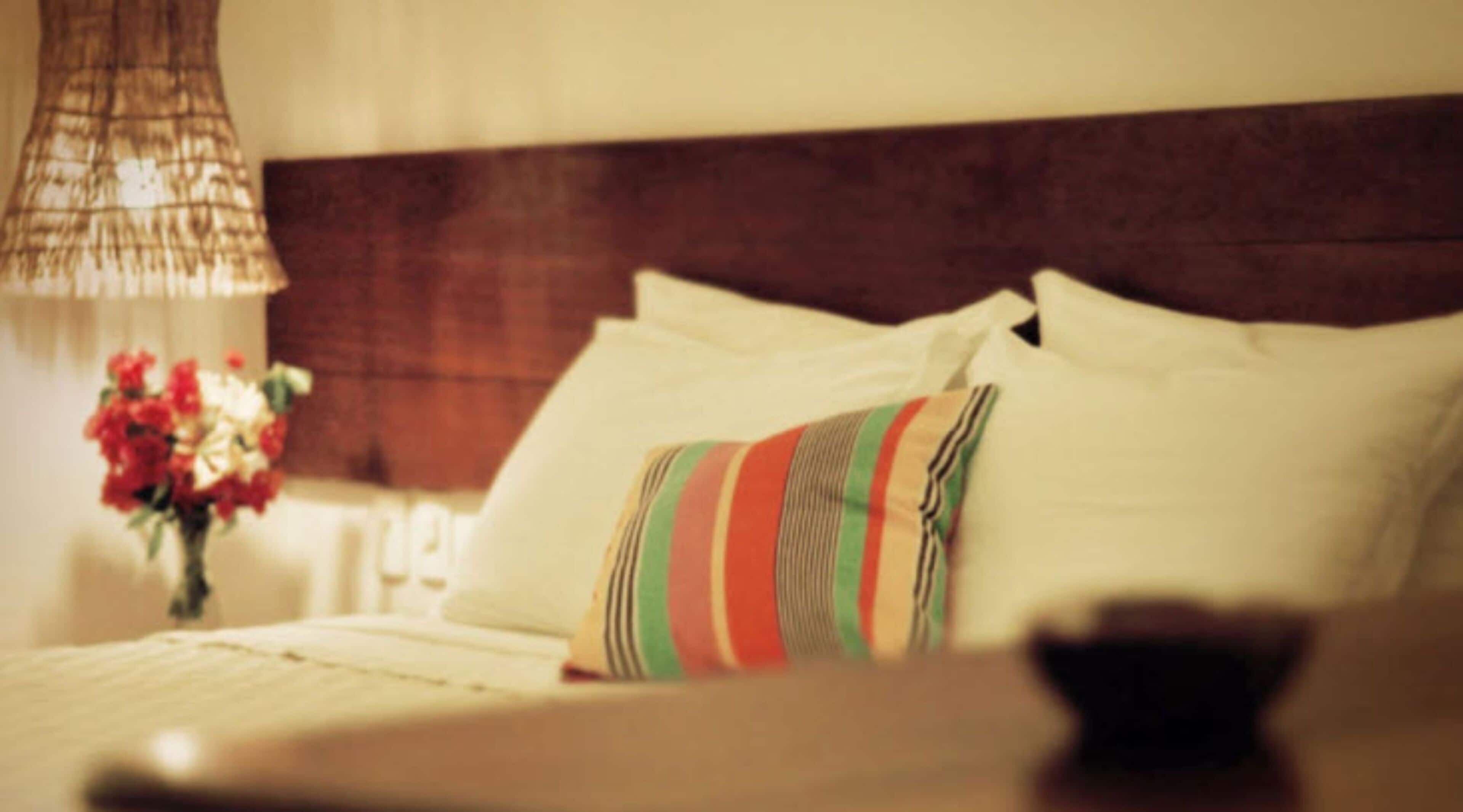 A bed with white pillows and a striped pillow, a vase with flowers, and a lamp.
