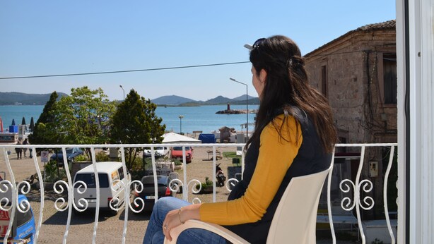 Una persona sentada en un balcón con vistas a la costa, con vehículos estacionados y edificios al fondo.