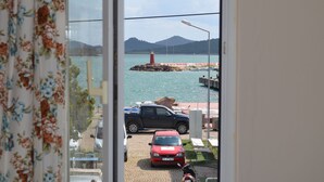 Vista desde una ventana con vista al puerto, un faro, vehículos estacionados y montañas al fondo.