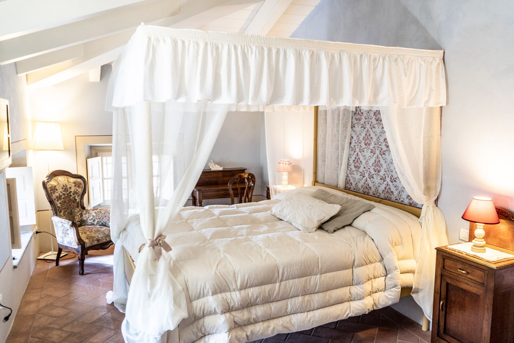 A bedroom with a four-poster bed, a chair, a nightstand, and a lamp.