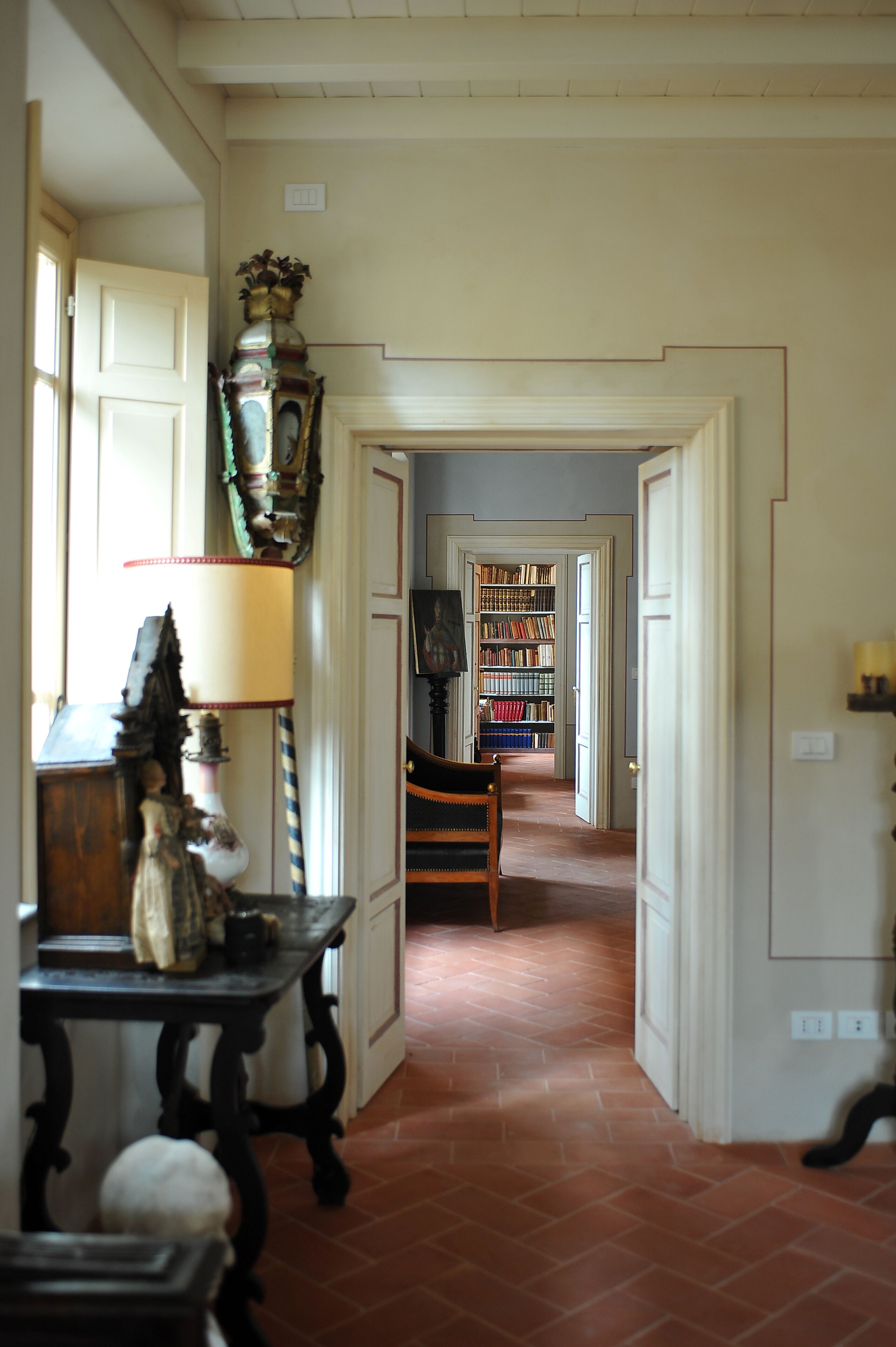 A hallway with a tiled floor, a wooden table with a lamp, a chair, and a bookshelf in the distance.