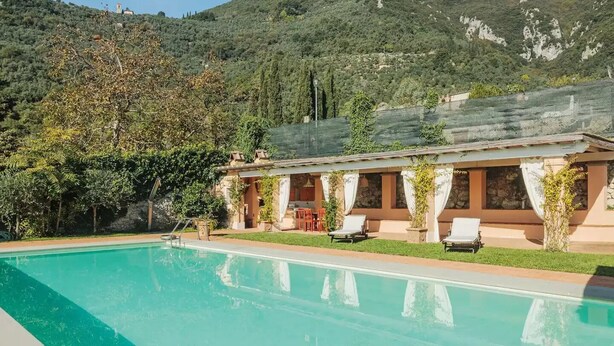 Zona junto a la piscina con un edificio, mobiliario exterior y montañas al fondo.