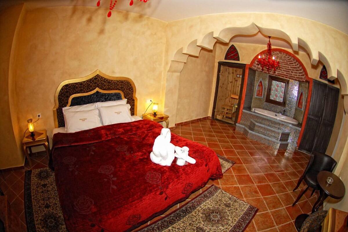 A bedroom with a large bed, a wooden headboard, a red bedspread, and a small table with a lamp.