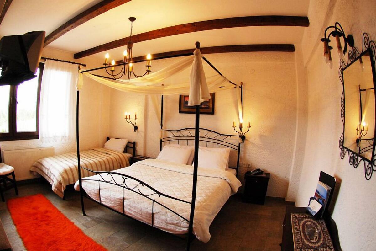 A four-poster bed with a canopy, two bedside tables, a chair, and a red rug in a room with white walls and wooden beams.