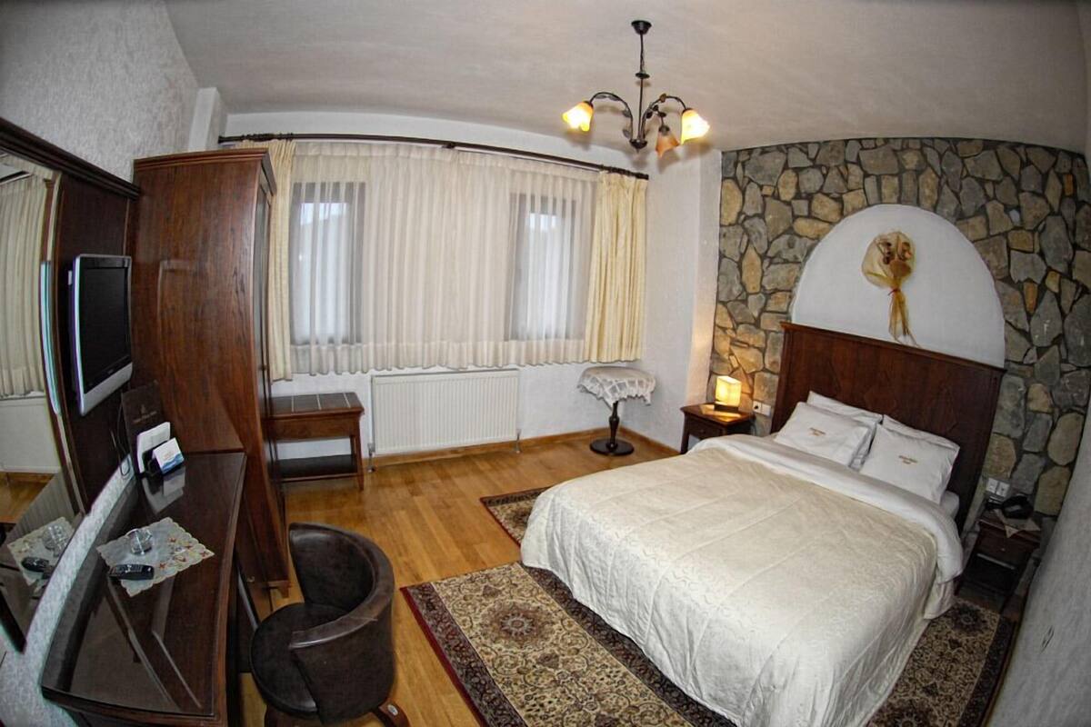 A bedroom with a stone wall, a bed with white bedding, a wooden wardrobe, a desk with a chair, a television, and a window with curtains.