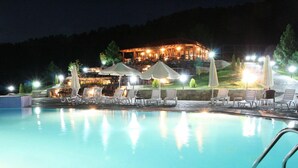 Área de piscina à noite com espreguiçadeiras e guarda-sóis, um edifício iluminado ao fundo e vista clara da piscina.