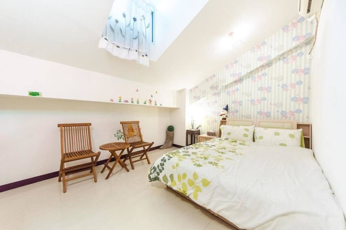 A bedroom with a bed, two wooden chairs, a small table, a nightstand, and a window with curtains.
