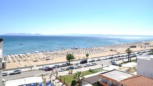 Une plage bordée de rangées de transats et de parasols, un parking et une vue sur la mer et les montagnes au loin.