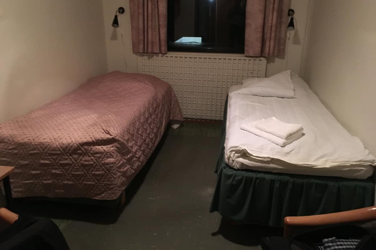 Una habitación pequeña y tenuemente iluminada con dos camas individuales, una con una colcha estampada y la otra con una colcha blanca lisa. Hay un radiador debajo de la ventana, y las paredes son lisas con un interruptor de luz y un aplique de pared.