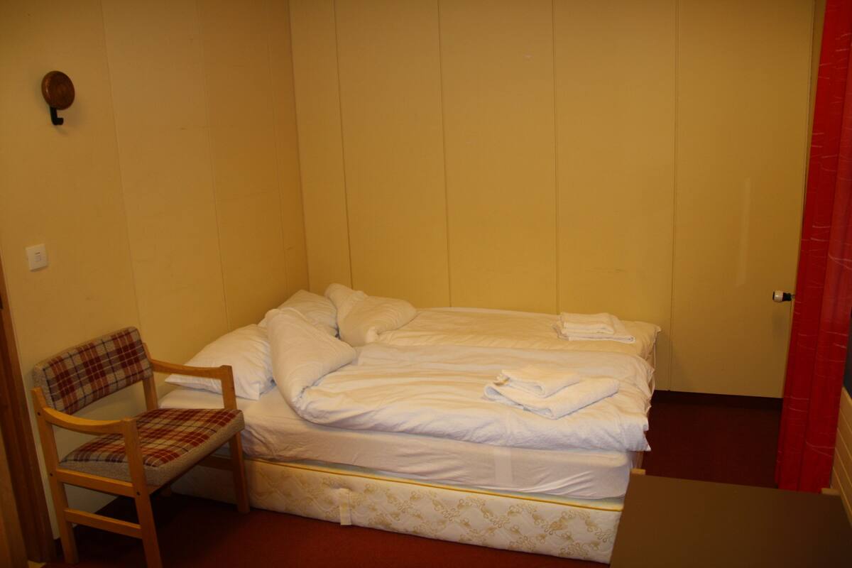 Una cama individual con sábanas blancas, una silla de madera con un cojín escocés y una cortina roja.