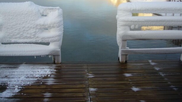 Eine Holzdielen-Terrasse mit mit Schnee bedeckten Stühlen, die auf einen zugefrorenen Kanal blicken.