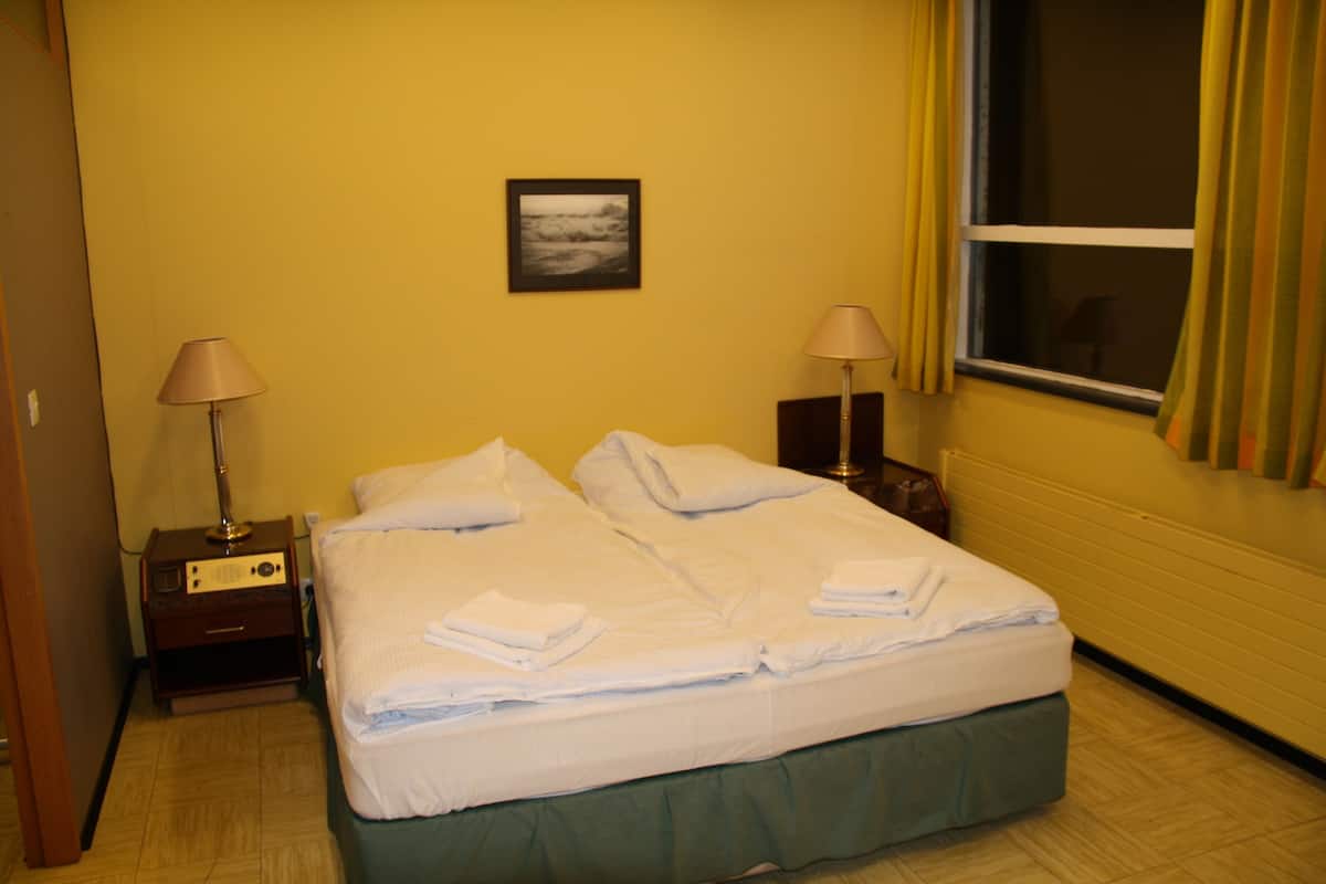 Habitación de hotel con cama, dos lámparas de noche, mesita y ventana con cortinas.