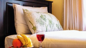 A neatly made bed with a white and green patterned pillow, a fruit bowl with bananas and an orange, and a glass of red wine on a bedside table. - Fedha Residences (Nairobi)