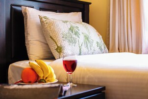 Una cama bien hecha con una almohada de blanco y verde estampado, un frutero con plátanos y una naranja, y una copa de vino tinto sobre una mesita de noche.