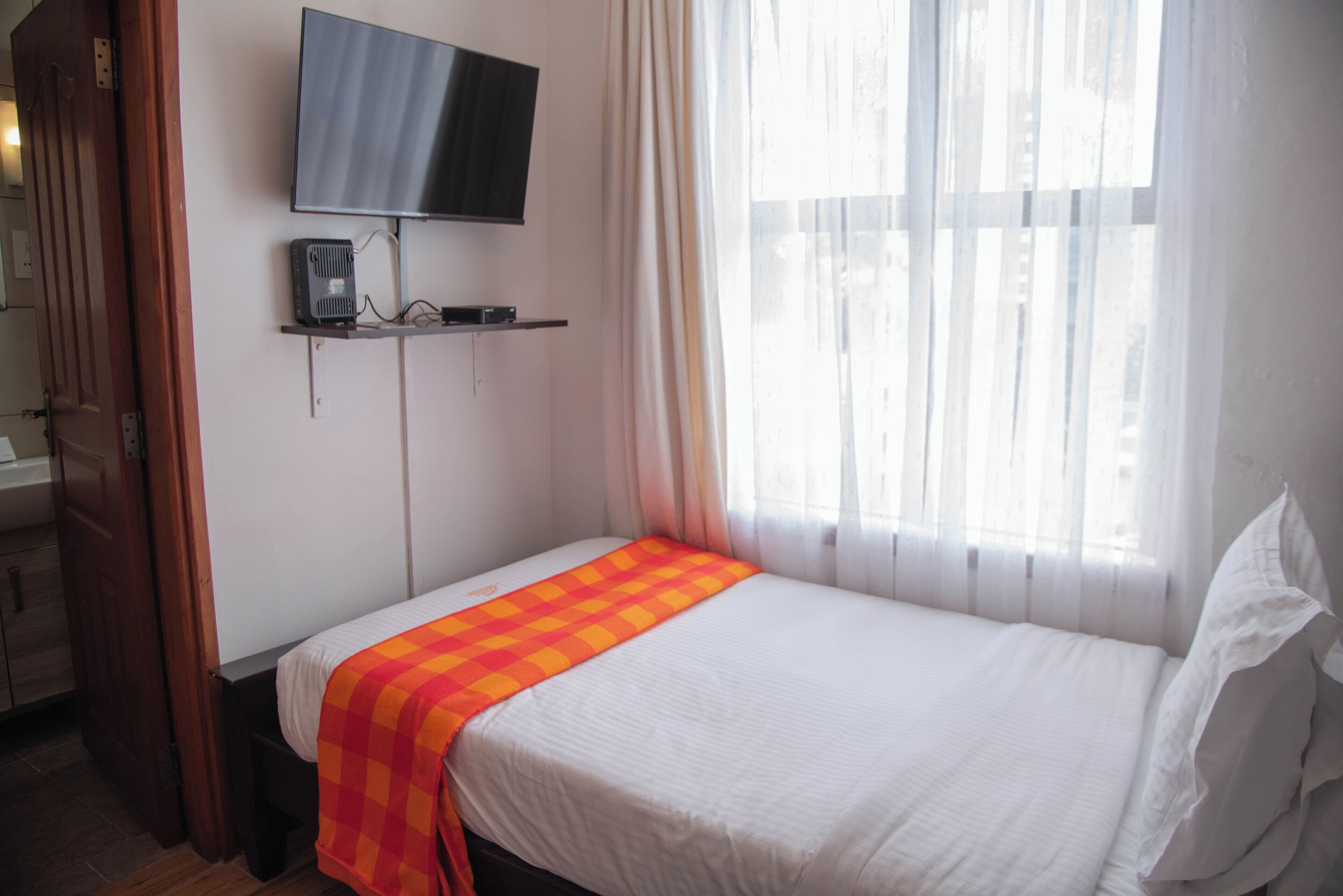 Un lit simple avec une couverture blanche et orange, un téléviseur à écran plat fixé au mur et une fenêtre ornée de rideaux légers.