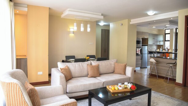 A living room with a sofa, armchairs, a coffee table with fruit and a vase, and a kitchen in the background. - Fedha Residences (Nairobi)