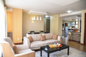Una sala de estar con sofá, sillones, una mesa de centro con frutas y un jarrón, y una cocina al fondo.