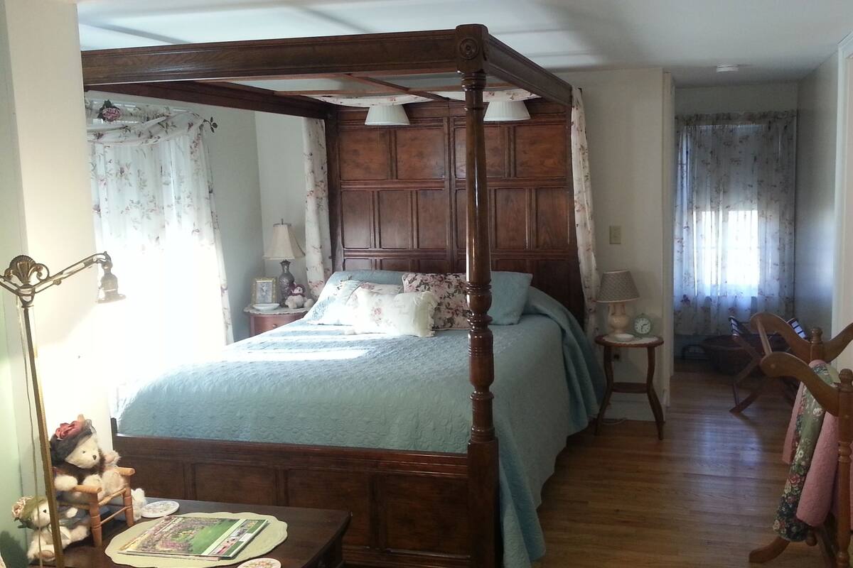 Een hemelbed met houten hoofdeinde, een nachtkastje met een lamp, een ander nachtkastje met een klok, en een stoel met een bloemenprint gordijn.