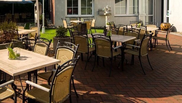 An outdoor patio with tables and chairs, a brick floor, and a building in the background. - Landhotel Elkemann (Ahaus)