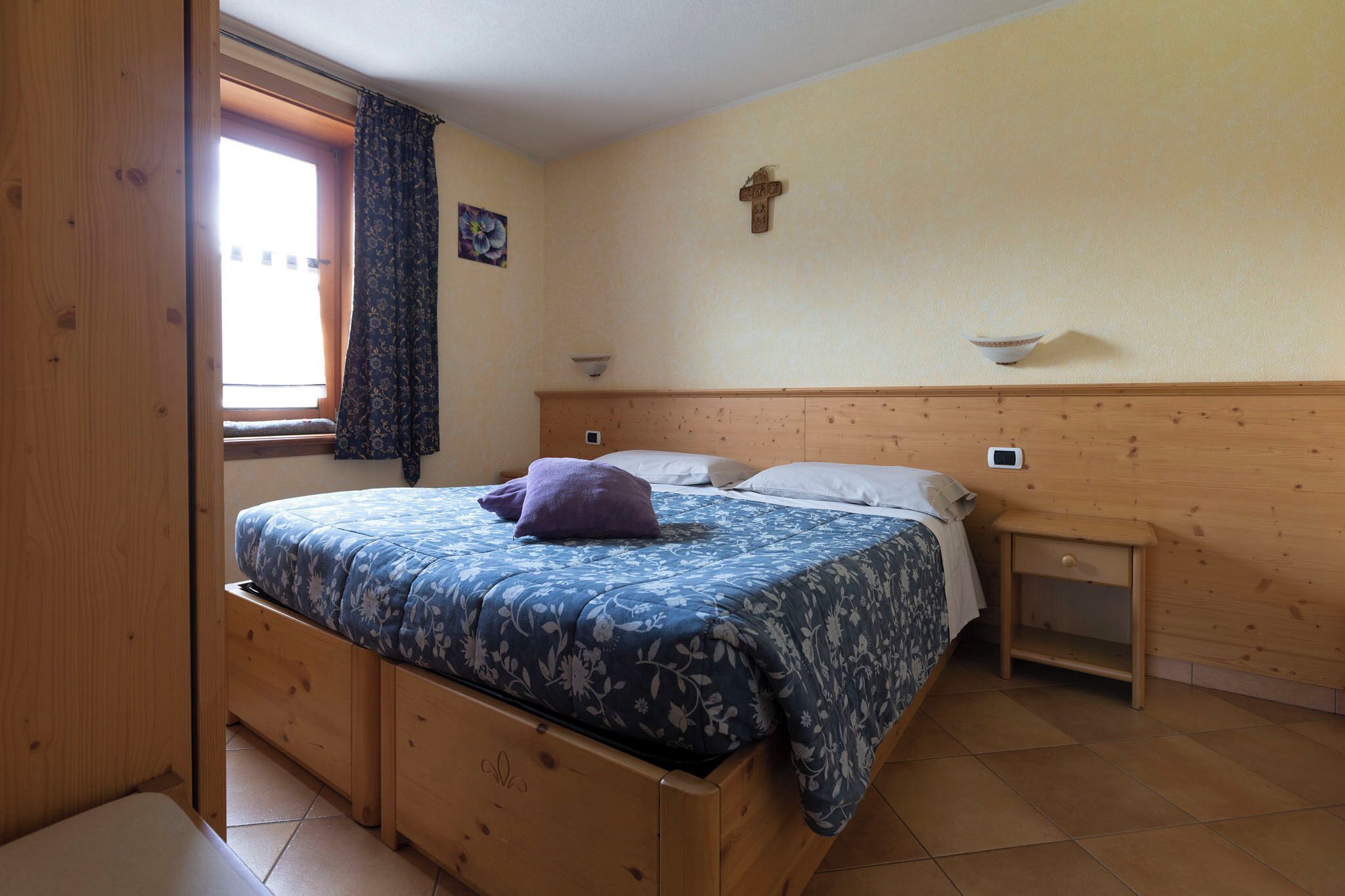 Un dormitorio con cama de madera, una mesita de noche, una lámpara de pared, una ventana con cortinas y una cruz en la pared.
