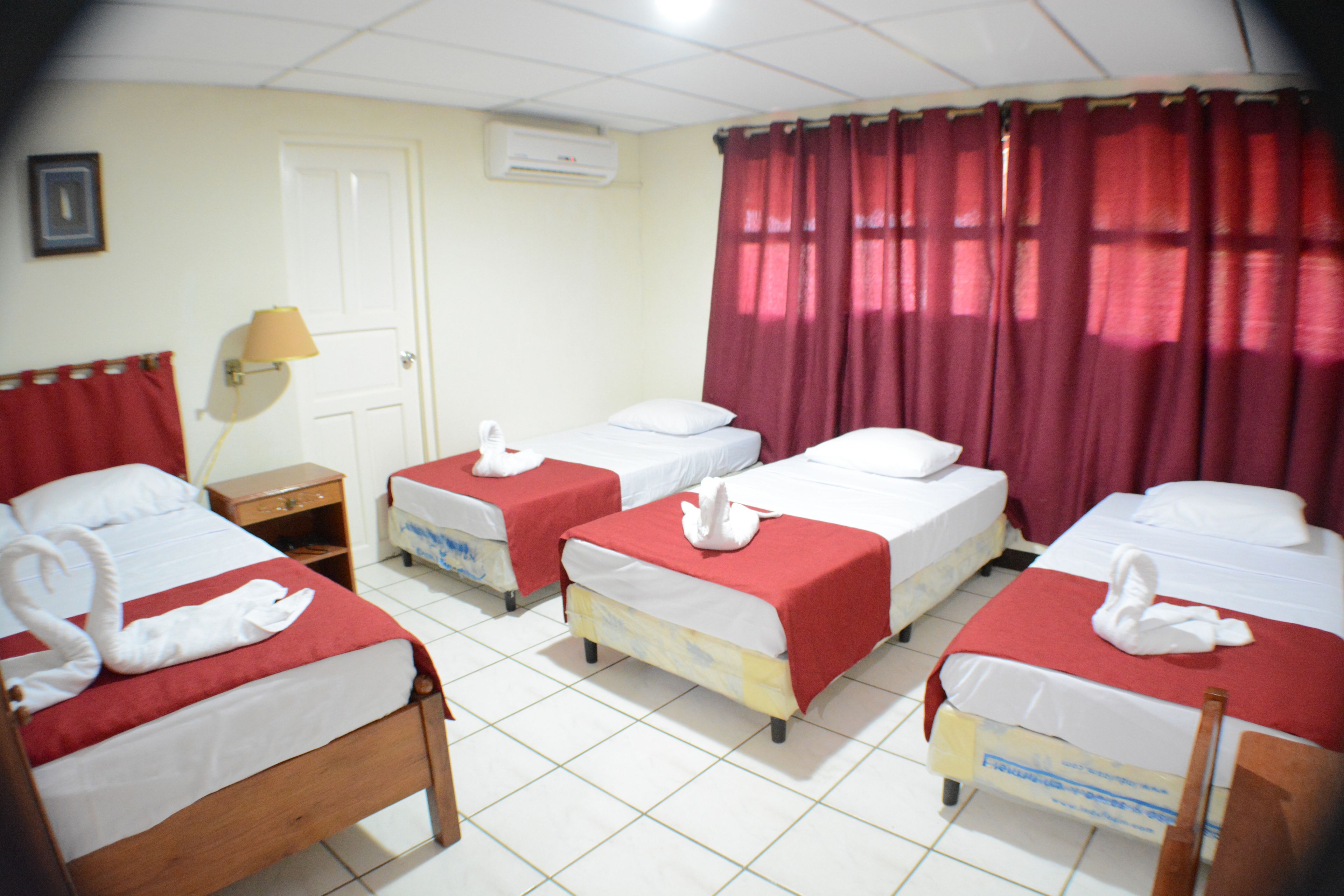 Habitación de hotel con cuatro camas, cada una con una manta roja y una toalla de ganso blanco. Hay una puerta blanca, una lámpara en una mesita de noche y una ventana con cortinas rojas.
