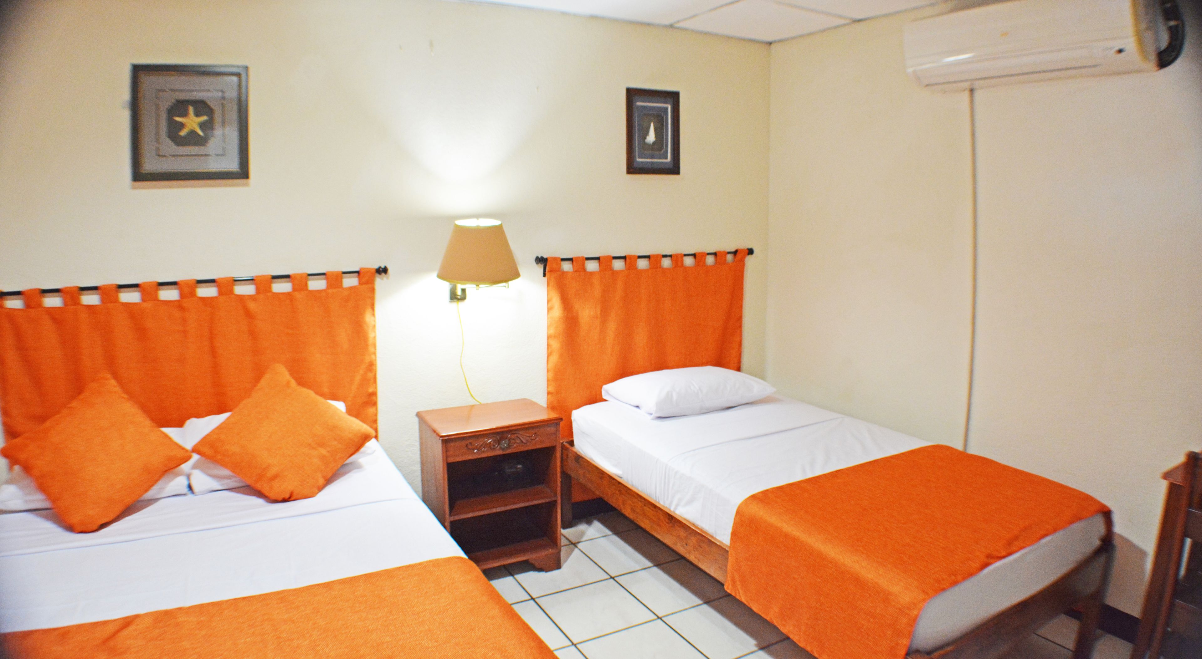 Habitación de hotel con dos camas, cada una con ropa de cama y almohadas anaranjadas, una mesita de noche, una lámpara y un aire acondicionado de pared.