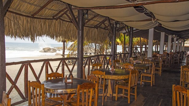 An outdoor restaurant with wooden tables and chairs, a thatched roof, and a view of the beach and ocean. - Hotel Suyapa Beach (León)