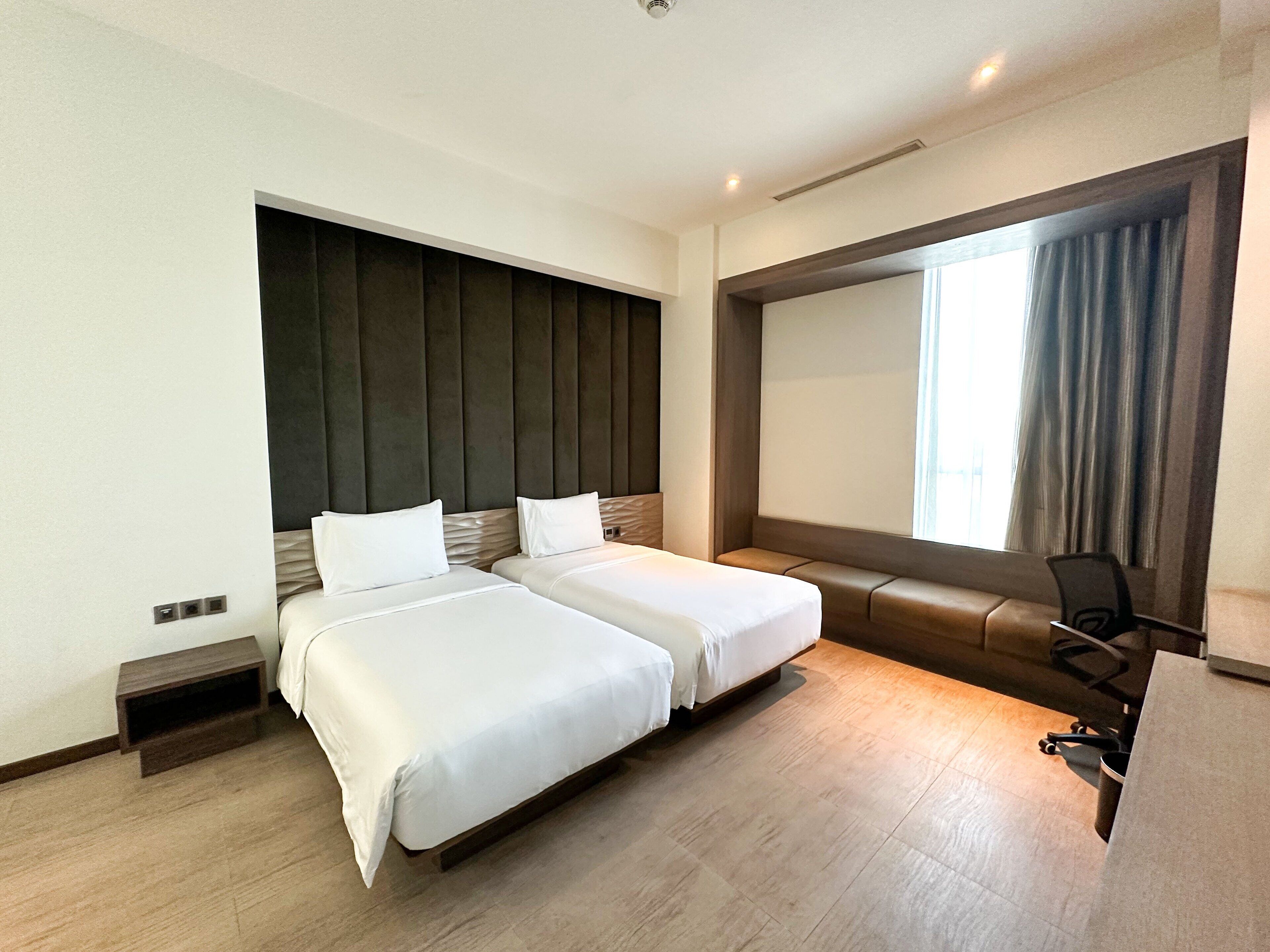 Hotelkamer met twee bedden, een bureau, een stoel, een groot raam met gordijnen en een ingebouwde televisie.