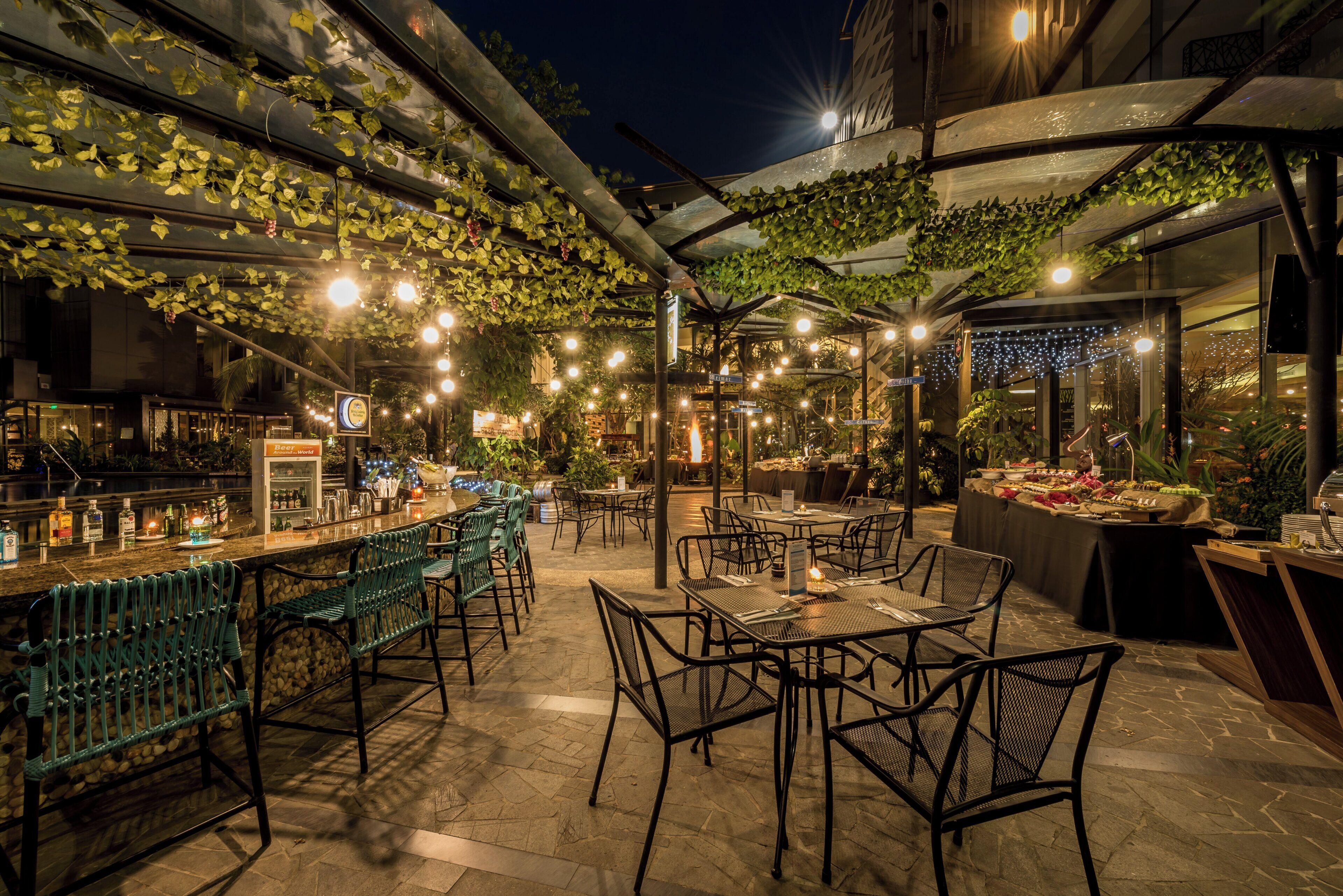 An outdoor dining area with tables and chairs, a bar with bottles, and greenery overhead.