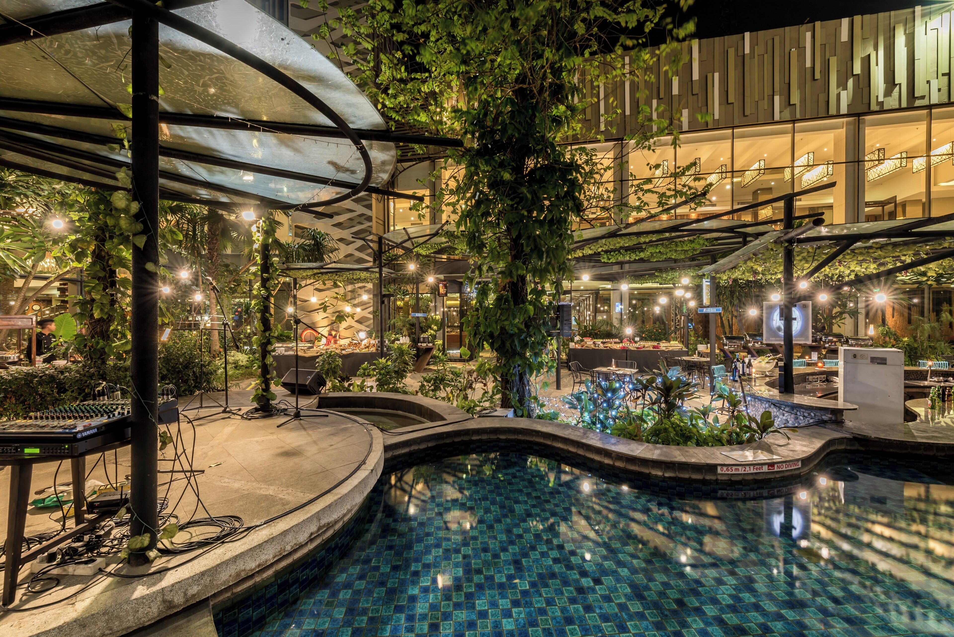 Un área de piscina interior con techo de vidrio, rodeada de follaje y mobiliario moderno.