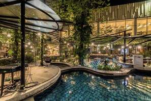 Un área de piscina interior con techo de vidrio, rodeada de follaje y mobiliario moderno.