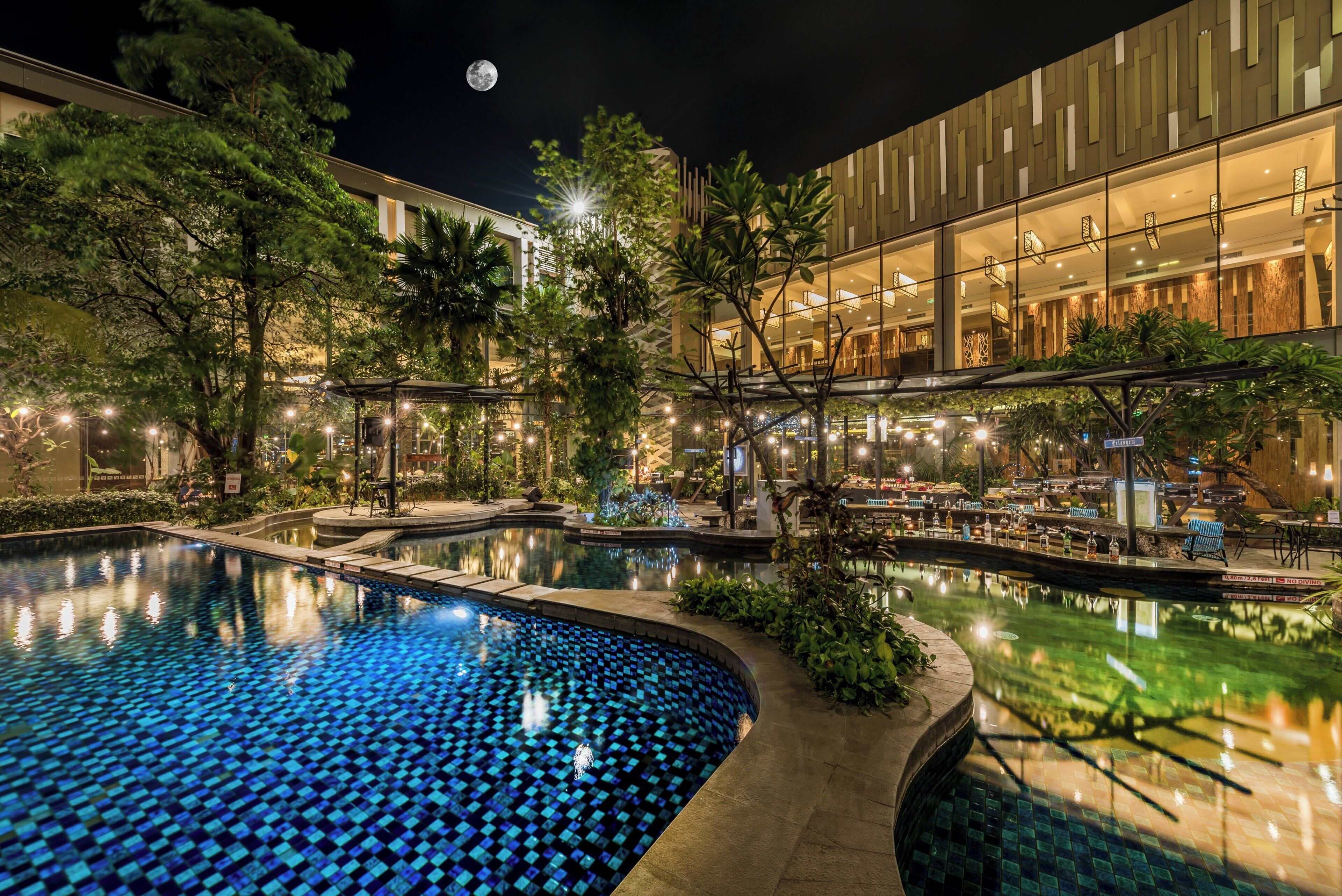 A nighttime pool area with a crescent moon in the sky, surrounded by trees and a building with a modern design.
