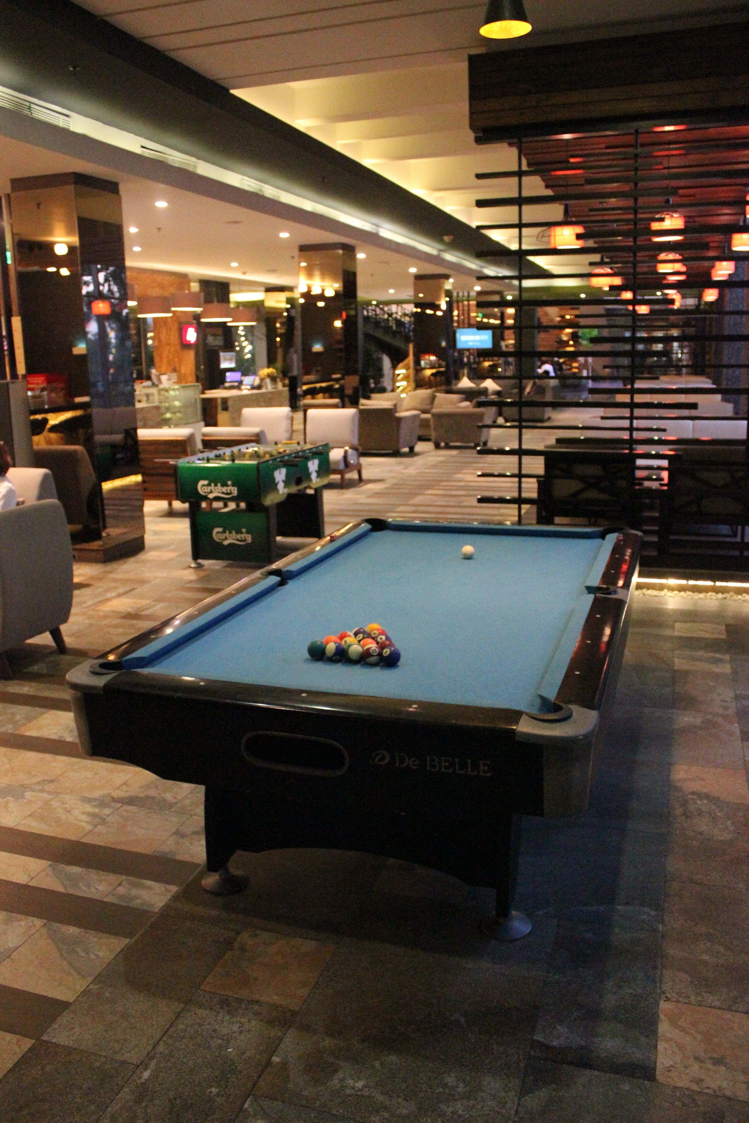 A pool table with balls, a billiard cue, and a pool table branded "De BELLE" in a hotel lobby with seating and a bar area.