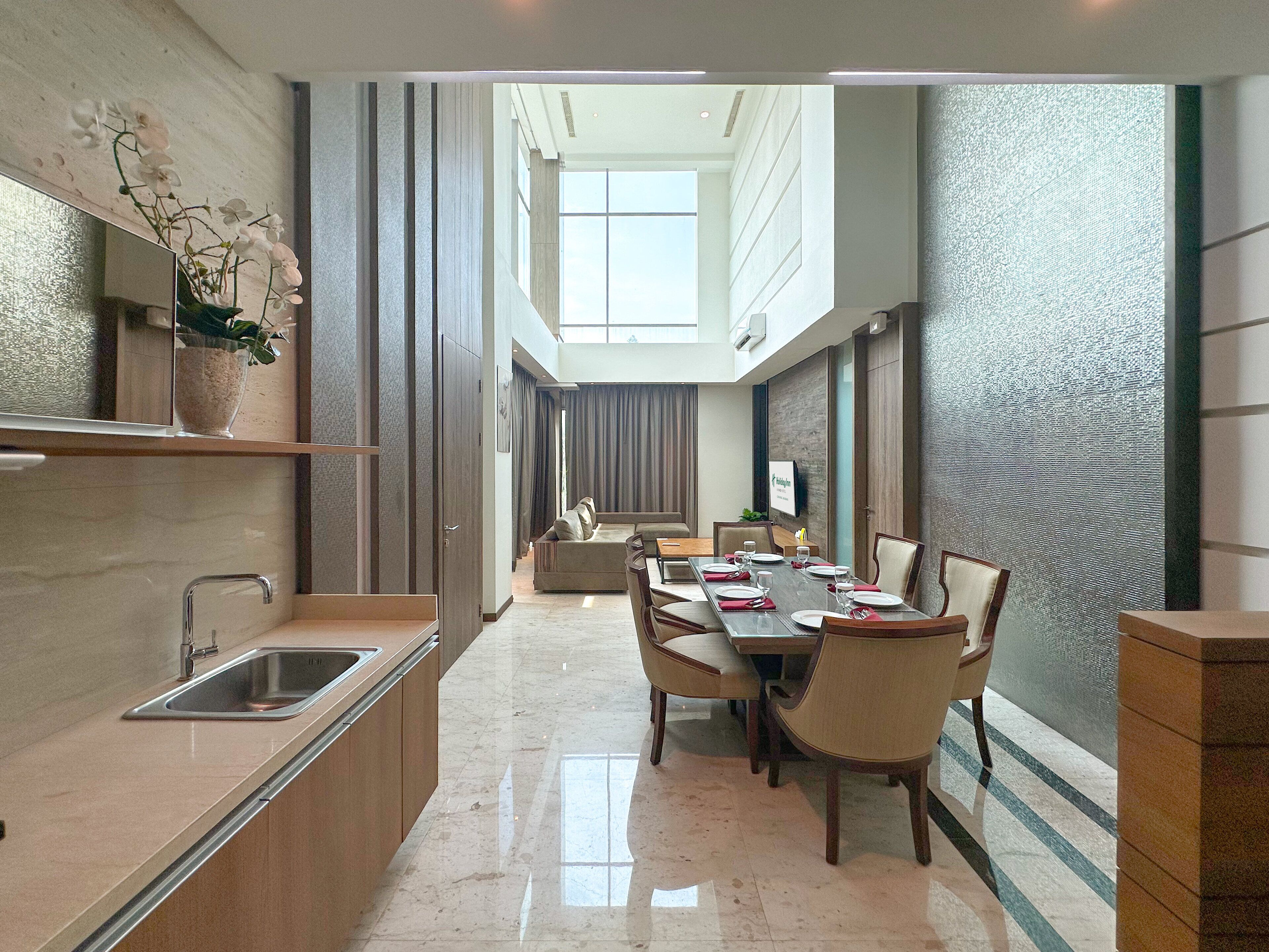 Un comedor moderno de hotel con una larga mesa dispuesta para una comida, una zona de cocina con un lavacabezas y una planta decorativa.