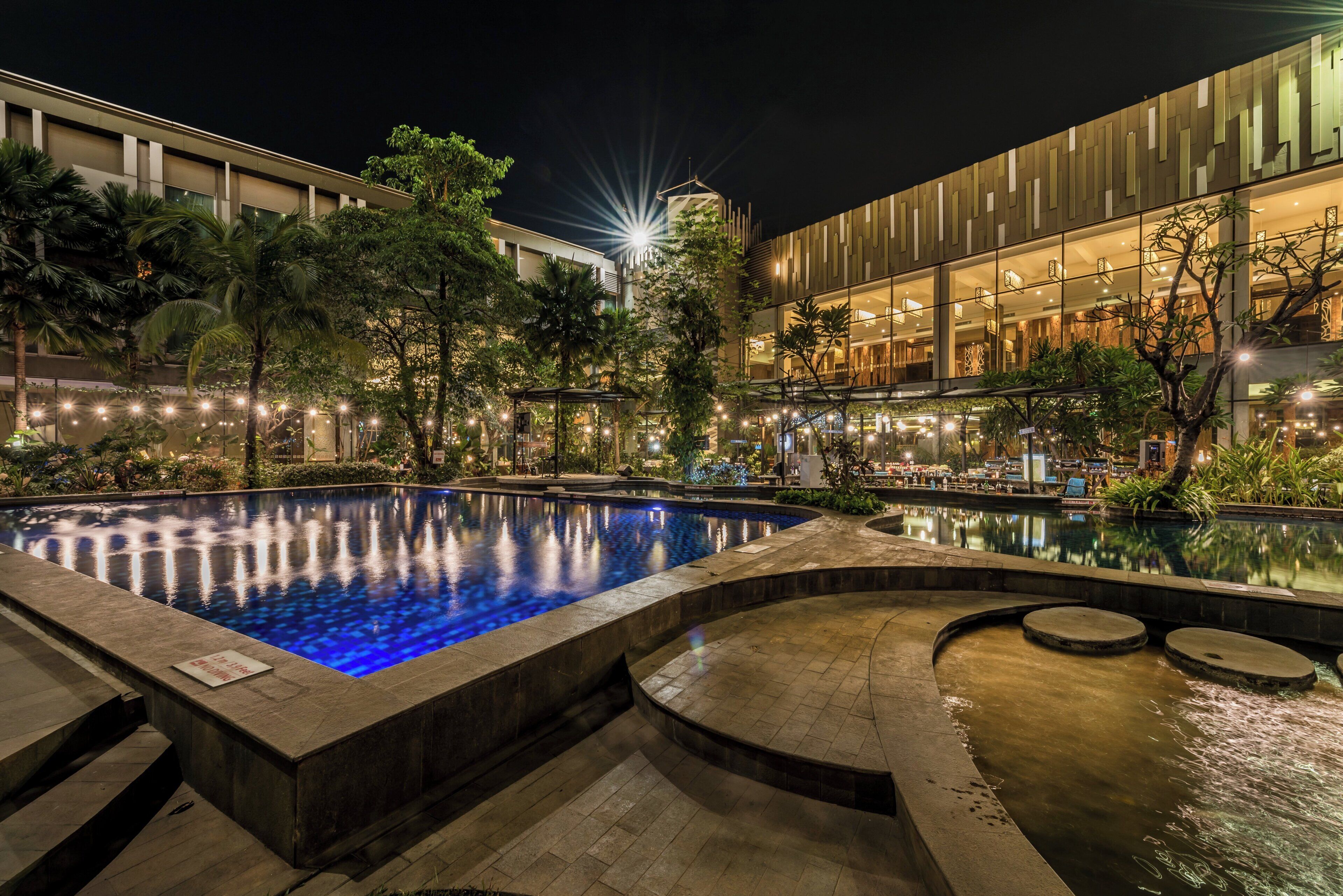 Un área de piscina nocturna con un edificio moderno al fondo, rodeada de árboles e iluminación ambiental.