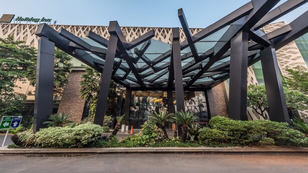 The exterior of a Holiday Inn with a modern glass and steel entrance, surrounded by lush greenery. - Holiday Inn Cikarang Jababeka by IHG (Cikarang)