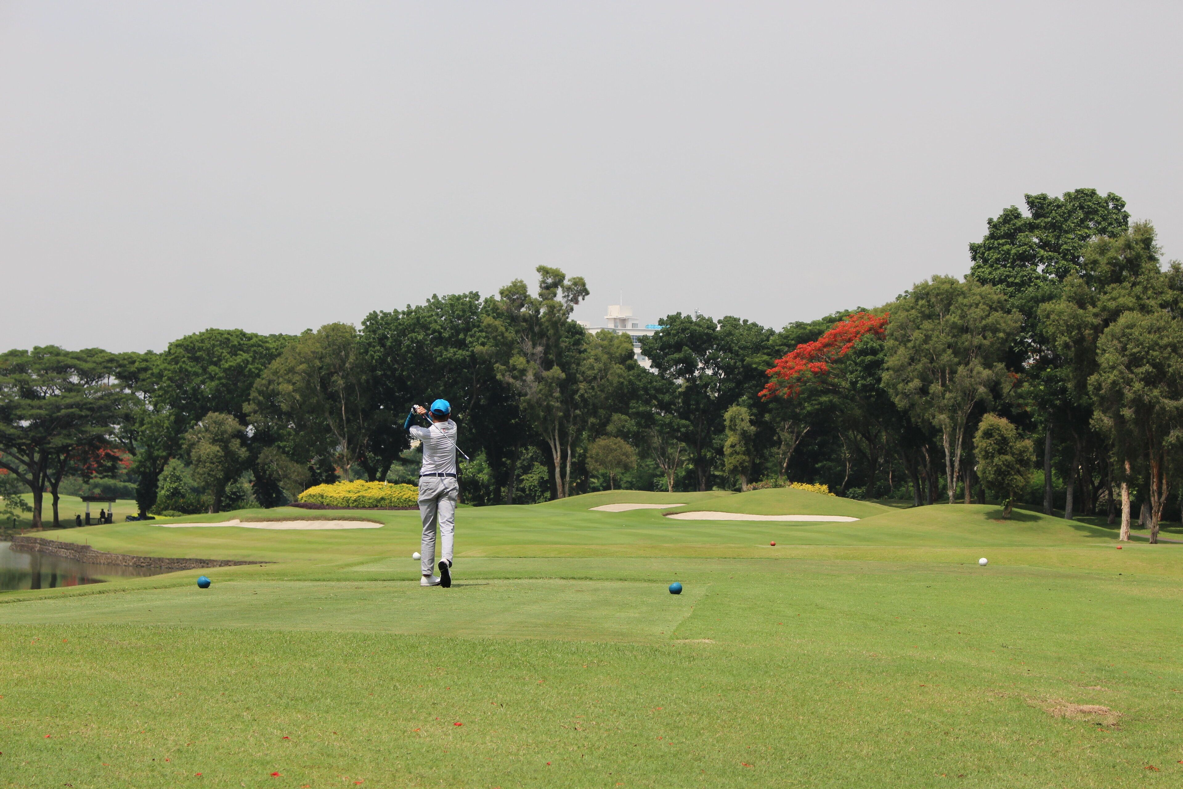 A golf course with a player in mid-swing, trees, and a distant building.