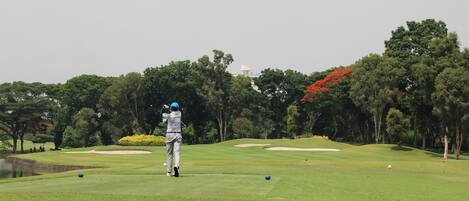A golf course with a player in mid-swing, trees, and a distant building.