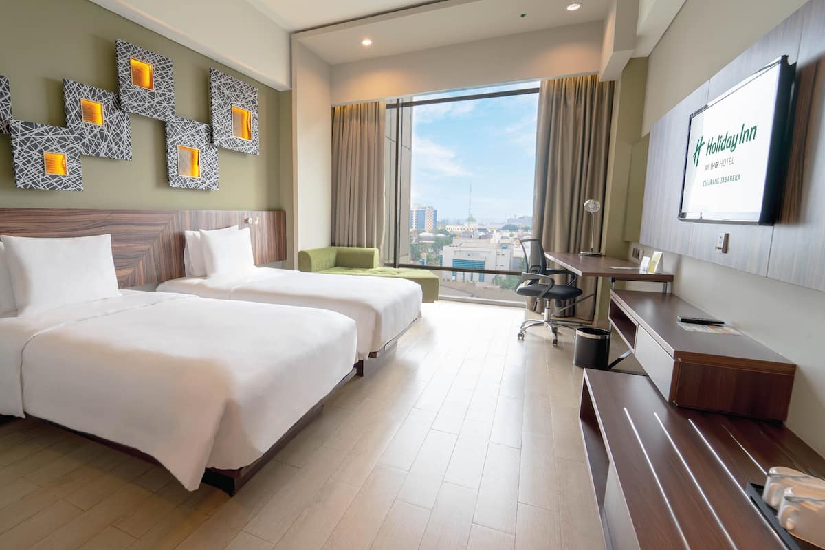A hotel room with two beds, a desk, a chair, a TV, and a view of the city.