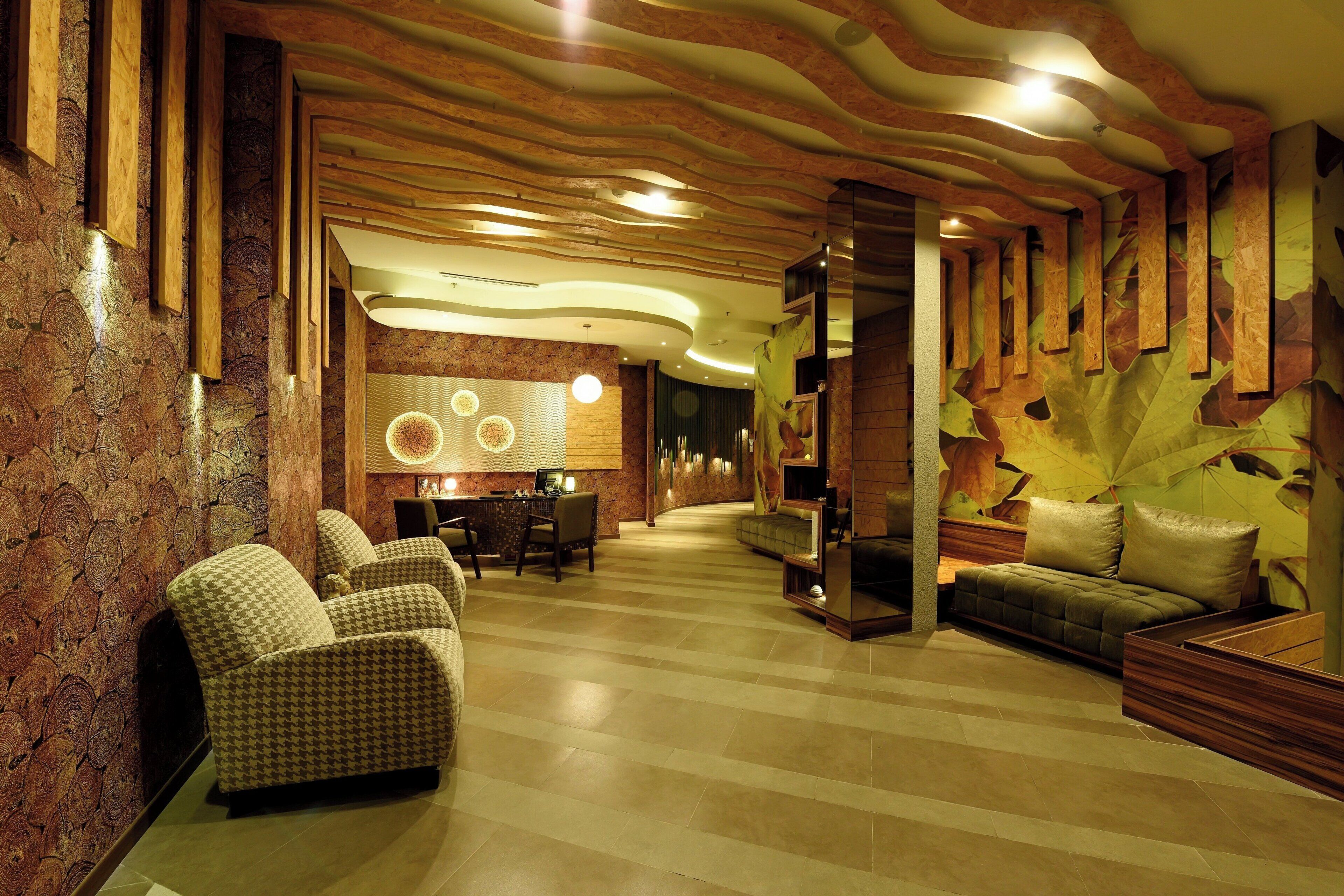 A spacious lobby with a modern design, featuring a central seating area with sofas and armchairs, a dining table, and a decorative wall with leaf patterns.