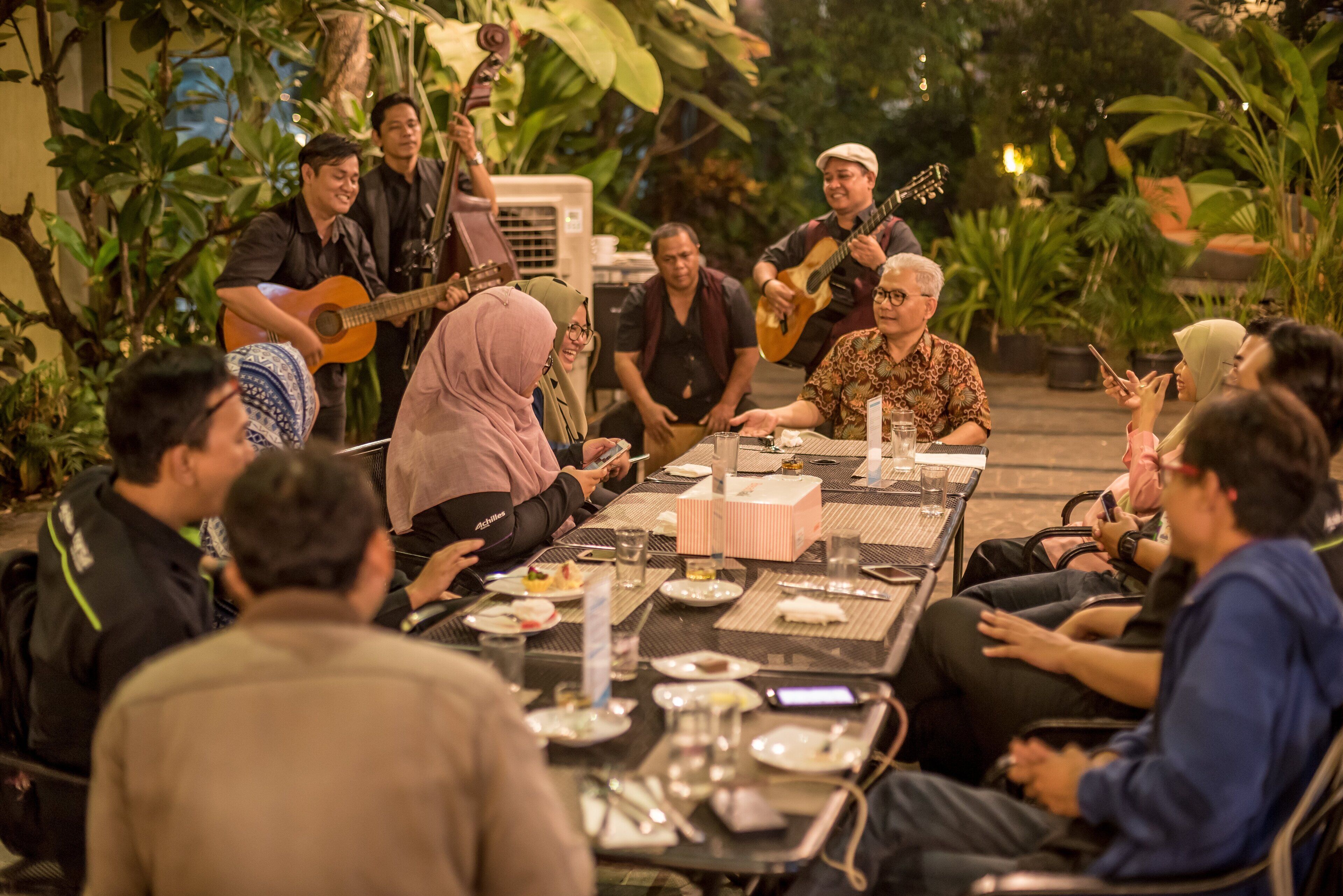 A group of people seated around a table outdoors with a band playing in the background.