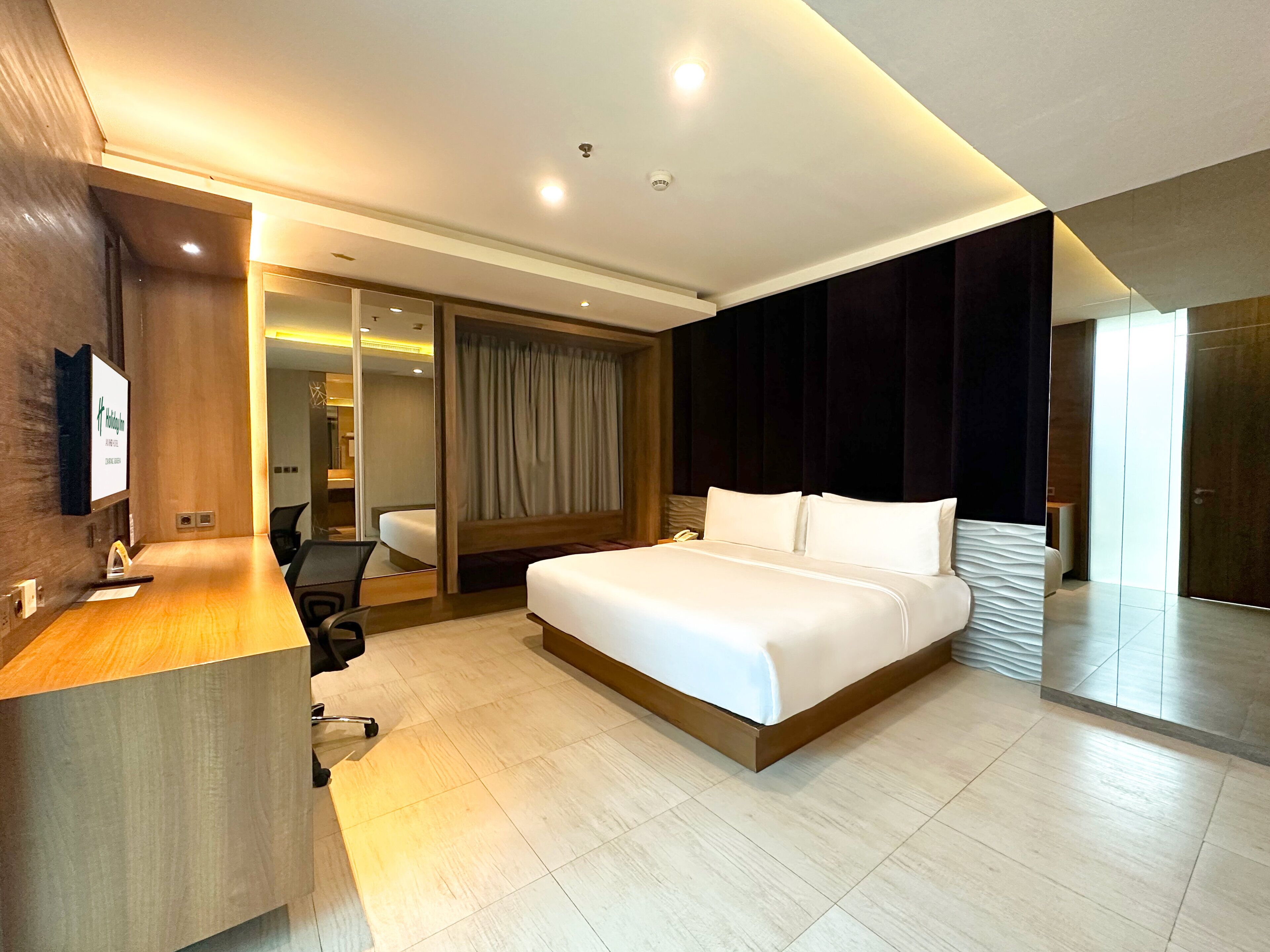 A modern hotel room with a large bed, a desk with a chair, a mirror, and a sliding door leading to a bathroom.