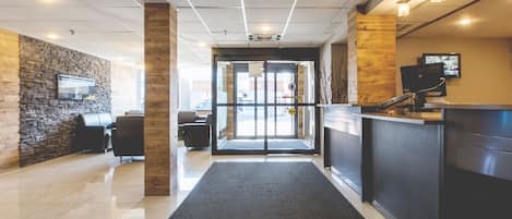 A hotel reception area with a modern design, featuring a reception desk, a seating area with chairs, a stone wall, and a glass door entrance.