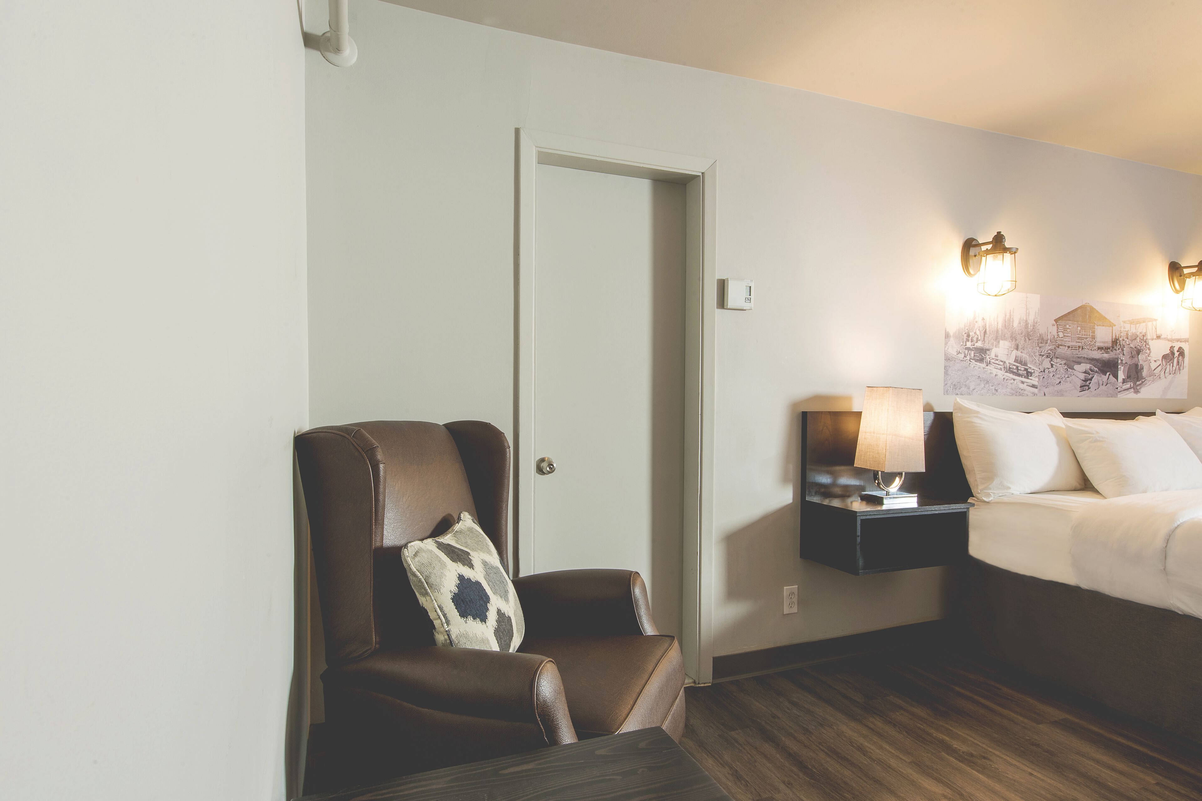 Una habitación de hotel con una butaca de cuero marrón, una mesita de noche con lámpara, una cama con ropa blanca y un aplique de pared.