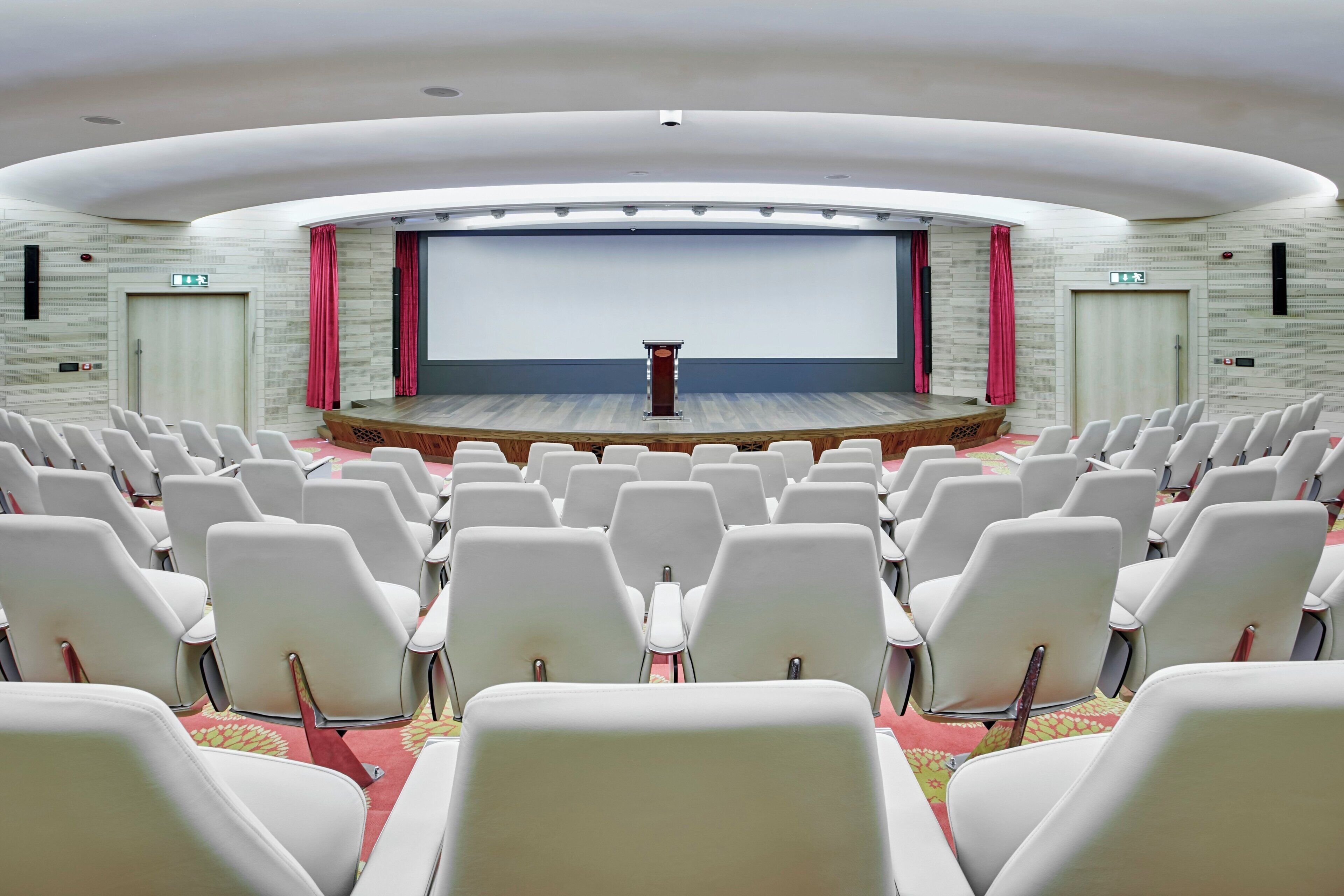 A conference room with a stage, rows of white chairs, a large screen, and a podium.